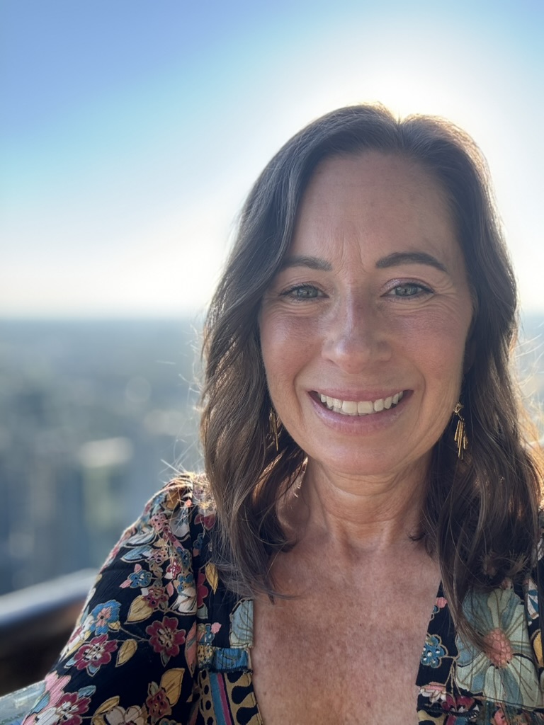 Carrie Beals smiling warmly outdoors, sky and horizon behind her, sun glowing at her shoulder.